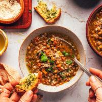 Holding a slice of garlic bread in one hand and a spoon in the other in a bowl of Italian lentil soup